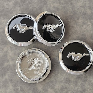 Puede incluir: Cuatro tapacubos de coche plateados y negros con un logotipo de caballo cromado. Los tapacubos son redondos y tienen un centro negro con un borde plateado.