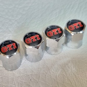 Könnte beinhalten: Vier verchromte Ventilkappen mit einem schwarzen und roten GTI-Logo. Die Kappen sind sechseckig und liegen auf einer hellen Oberfläche. Das GTI-Logo ist in roten Buchstaben gehalten.