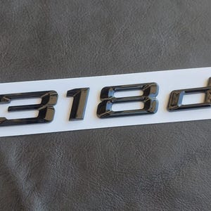 May include: Glossy black "318d" emblem, likely for a vehicle, is displayed on a white strip against a textured dark gray background. The numbers and letter are bold and modern in style, suggesting a car part or accessory.
