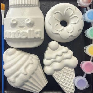 May include: A paint-your-own kit with a Nutella jar, donut, and two ice cream cone shapes. Includes a paintbrush and a set of paint pots with various colours. The items are white and ready to be painted.