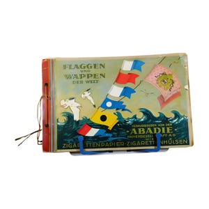 Puede incluir: Un libro vintage titulado "Flaggen und Wappen der Welt" (Banderas y Escudos de Armas del Mundo) con una ilustración colorida de banderas y una gaviota en la portada. El libro está publicado por Abadie Papiergesellschaft A.G. y presenta una bandera azul, roja y blanca con un círculo amarillo en el centro.