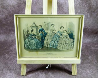 Könnte beinhalten: Ein gerahmter Vintage-Druck von Frauen in aufwendigen Kleidern, wahrscheinlich aus dem 19. Jahrhundert, auf einer kleinen Holzstaffelei ausgestellt. Der Druck zeigt eine Szene mit mehreren Figuren in historischer Kleidung, mit einer weichen, gedämpften Farbpalette.