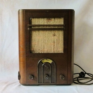 Könnte beinhalten: Ein Vintage-Holzradio mit einem braunen Holzkasten und einem Lautsprechergitter aus Stoff. Das Radio hat ein Zifferblatt mit einem Zeiger und zwei Knöpfe an der Vorderseite.