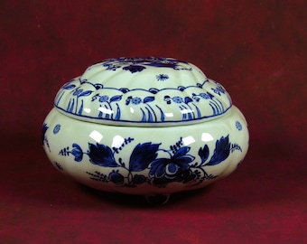 May include: A white ceramic lidded jar with blue and white floral designs. The jar has a rounded shape and sits on three small feet.