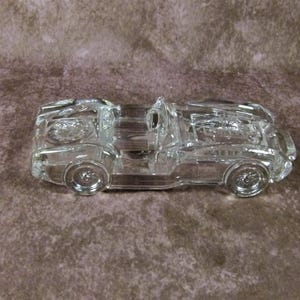 May include: A clear glass figurine of a vintage sports car. The car is detailed with a windscreen, wheels, and a driver's seat.