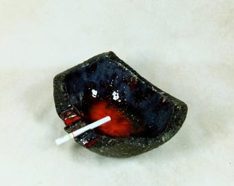 Puede incluir: Un cenicero de cerámica con un esmalte azul oscuro y rojo. Un cigarrillo está apoyado en el cenicero.