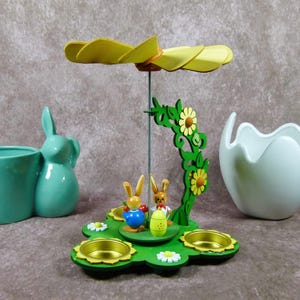 Könnte beinhalten: Eine hölzerne Osterpyramide mit gelbem und grünem Design. Die Pyramide hat zwei Hasen, Blumen und einen drehenden Propeller oben. Es gibt vier Kerzenhalter auf der Basis.