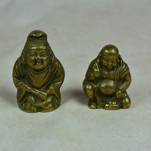 Könnte beinhalten: Zwei kleine, antike Buddha-Statuen aus Messing. Eine steht, trägt einen Hut und eine Robe und hält eine Schriftrolle. Die andere hockt, mit einem großen Bauch und einem ruhigen Ausdruck. Beide haben einen goldenen Farbton.
