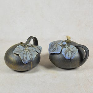 Peut inclure: Deux théières en céramique avec une finition métallique foncée. Chaque théière est dotée d'une anse et est ornée de feuilles et de baies bleu clair. Les théières ont un aspect unique et artisanal, suggérant qu'il s'agit d'objets décoratifs.