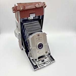 Puede incluir: Cámara Polaroid Land Camera modelo 350 vintage con parte superior de cuero marrón y cuerpo plateado. La cámara está abierta, revelando el fuelle y la lente. Sobre una superficie blanca.