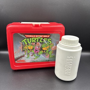 以下が含まれることがあります： 赤色のプラスチック製ランチボックスと、Teenage Mutant Ninja Turtlesのグラフィック、白い魔法瓶。ランチボックスには「Teenage Mutant Ninja Turtles」の文字があり、魔法瓶には「Thermos」の文字が印刷されています。