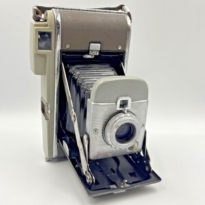 May include: Vintage Polaroid camera with a gray and brown leatherette body, silver accents, and a lens. The camera is open, revealing the bellows and lens. The word "Polaroid" is printed on the front.