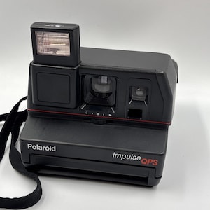 Puede incluir: Cámara instantánea Polaroid Impulse QPS negra con flash incorporado y correa para el cuello negra. La cámara tiene una línea roja y los logotipos de Polaroid e Impulse QPS son visibles.