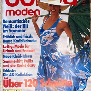 Puede incluir: Una portada de revista con una mujer que lleva un vestido estampado en azul y blanco y un sombrero de paja. La revista se titula "Burda Moden" y tiene el texto "Über 120 Schnitte" en la portada. La revista es de junio de 1985.