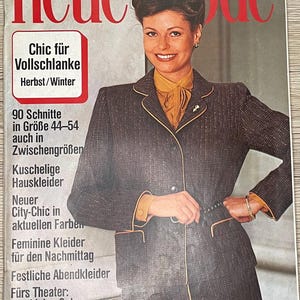 Può includere: Una copertina di rivista con una donna che indossa una giacca di tweed marrone e nero con un colletto giallo. La rivista si intitola "neue mode" e il testo "Chic für Vollschlanke Herbst/Winter" è visibile. La rivista è un'edizione speciale per la moda curvy.