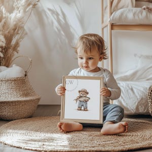 May include: A child holds a framed print of a brown teddy bear wearing a blue shirt and a hat, holding a fish. The print has a white background and a wooden frame.