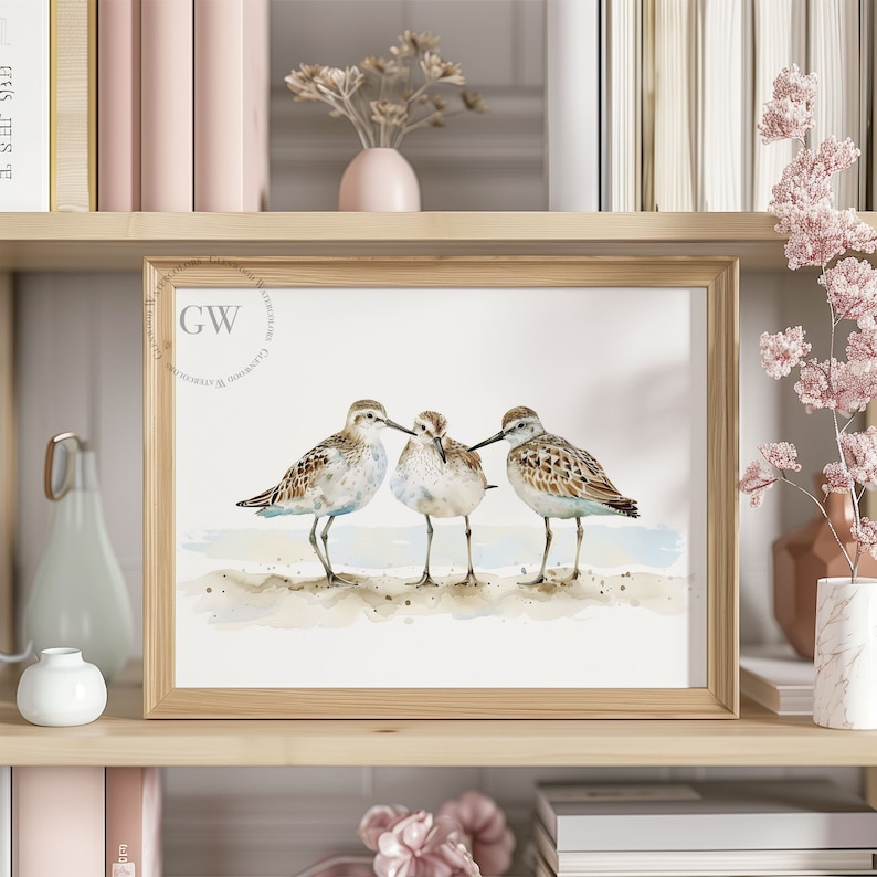 May include: A watercolor painting of three sandpipers standing on a sandy beach. The birds are brown and white with long legs and beaks. The painting is framed in a light wood frame.