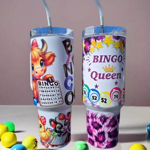 May include: Two white tumblers with lids and straws. The tumbler on the left has a cartoon cow and a bingo card. The tumbler on the right has a pink and purple leopard print design and the text "Bingo Queen" with a crown.