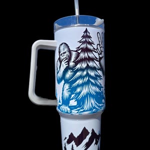 May include: White travel mug with a blue and black graphic of a Bigfoot holding a pine tree and making a peace sign. The bottom of the mug has a black and white graphic of mountains and trees.