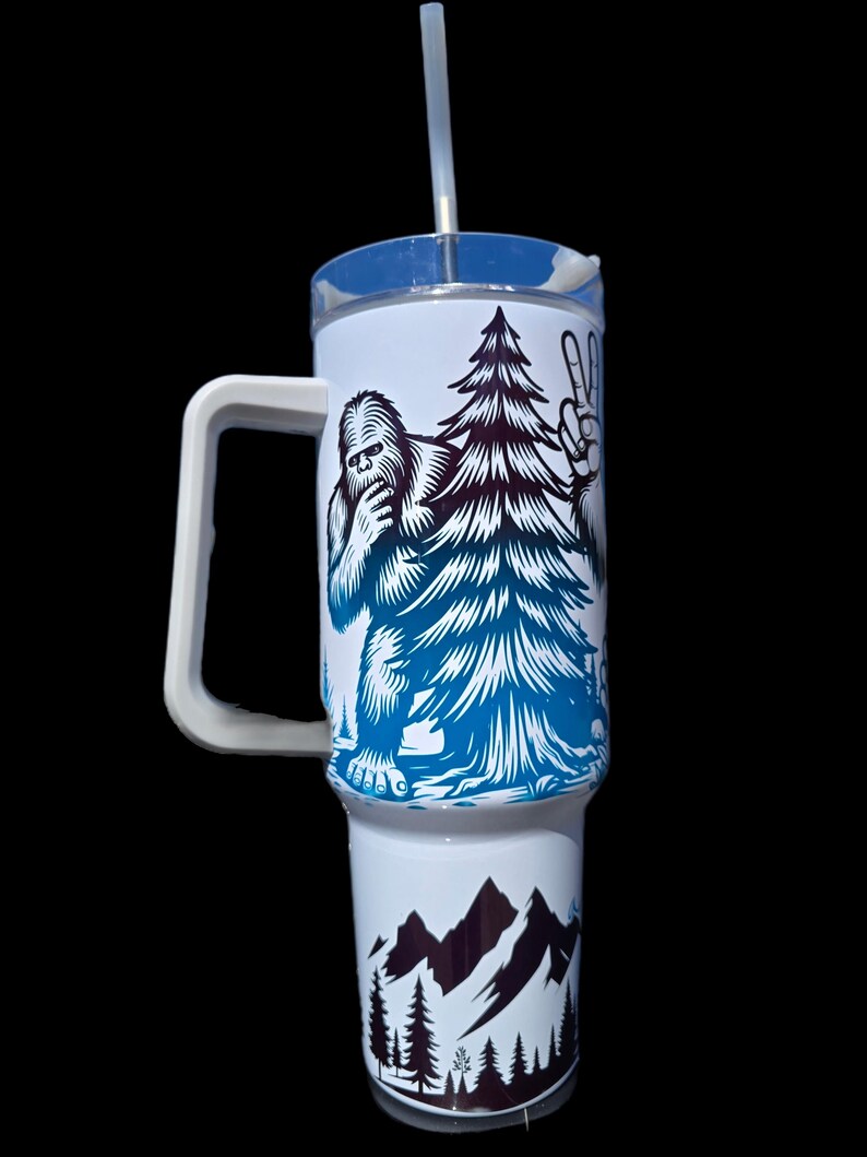 May include: White travel mug with a blue and black graphic of a Bigfoot holding a pine tree and making a peace sign. The bottom of the mug has a black and white graphic of mountains and trees.