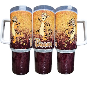 May include: Three insulated tumblers with a gold glitter background and a cartoon image of Tigger from Winnie the Pooh. The tumblers have a white lid and a brown bottom. The text "Tigger" is printed on the front of the tumblers.