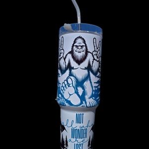May include: A white tumbler with a blue and black graphic of a Bigfoot with peace signs. The tumbler has a blue and white text that reads "Not all who wander are lost".