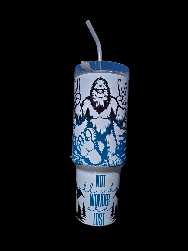 May include: A white tumbler with a blue and black graphic of a Bigfoot with peace signs. The tumbler has a blue and white text that reads "Not all who wander are lost".