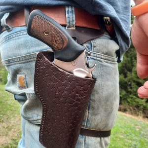 May include: A brown leather holster with a patterned design, holding a silver revolver with a wooden grip. The holster is worn on a person's hip, secured with a brown leather belt.
