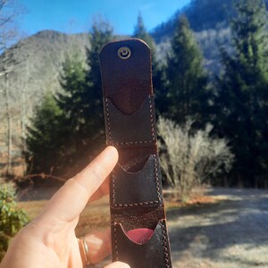 May include: A brown leather guitar pick holder with three slots. The holder has a decorative stitch around the edges and a small metal button on the top.