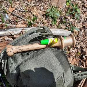 May include: A green and yellow fishing lure attached to a wooden fishing rod, resting on a green canvas backpack.