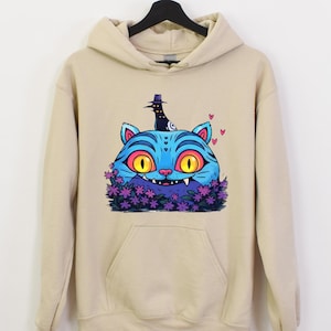 May include: A beige hoodie with a front pocket and a drawstring hood. The front features a vibrant cartoon illustration of a blue cat with yellow eyes, a black hat, and purple flowers. The cat design is whimsical and colorful.