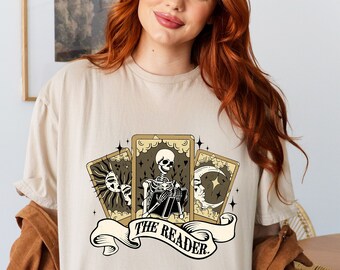 May include: A beige t-shirt with a graphic of three tarot cards featuring a skeleton holding a book. The cards are surrounded by stars and the text "THE READER." is printed below.