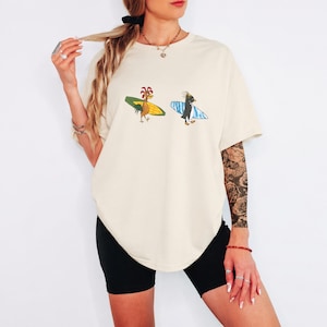 May include: An oversized cream-colored t-shirt featuring a graphic print of a cartoon rooster carrying a yellow surfboard and a cartoon penguin carrying a blue and white striped surfboard. The shirt has a crew neckline and short sleeves.