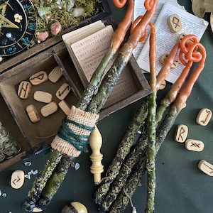 May include: A collection of handcrafted ritual candles with a rustic aesthetic. The candles feature a green base, transitioning to orange at the top, with a spiral design. Wooden runes, a compass, and a wooden box with cards are also visible.