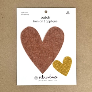 May include: Two iron-on patches in the shape of hearts. The larger heart is a reddish-brown color and the smaller heart is a mustard yellow. The text on the card reads "patch iron-on / applique" and "rebundance create more + waste less".