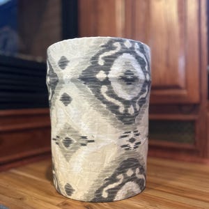 May include: Cylindrical decorative vase with a cream and gray ikat pattern. The vase is made of a textured material and sits on a wooden surface. The pattern features geometric shapes and organic forms in varying shades of gray.