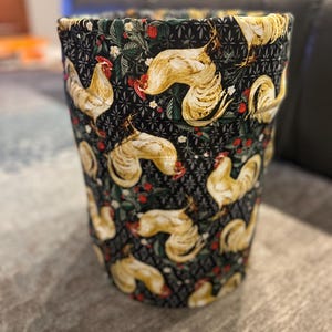 May include: A cylindrical fabric wastebasket featuring a black background with a repeating pattern of golden-yellow roosters, small red berries, and green leaves, accented by a subtle geometric star-like print throughout the dark fabric.