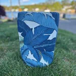 May include: A cylindrical container with a blue and white tropical leaf pattern. The fabric-covered item is set on a bed of green grass, with a blurred background of a body of water and trees.