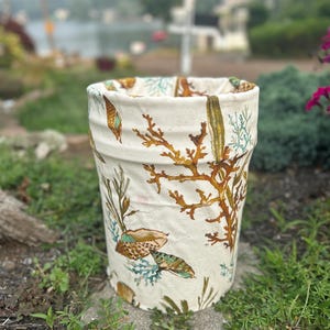Peut inclure: Un pot de fleurs cylindrique en céramique blanc cassé orné d'un imprimé botanique détaillé représentant des algues brunes, du corail et divers coquillages dans des tons sarcelle, marron et beige. Le récipient a une surface texturée.