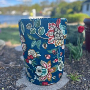 May include: A cylindrical planter with a dark teal background and a floral pattern in red, white, and teal. The planter is outdoors, sitting on a stone surface, with a blurred background of a lake and trees.