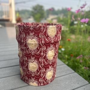Peut inclure: Un panier de rangement cylindrique en tissu avec un fond rouge foncé et un motif toile de Jouy crème répétitif. Le design se compose de cadres ovales ornés contenant des scènes pastorales détaillées, reliés par des volutes et des motifs floraux.