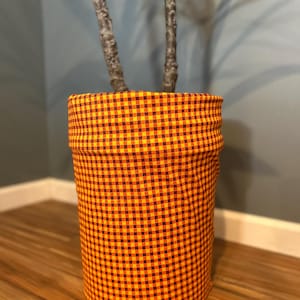 May include: A cylindrical plant pot cover with a yellow and burgundy checkered pattern. The fabric pot cover is wrapped around a plant with a brown trunk and green leaves. The pot cover is sitting on a wooden floor.