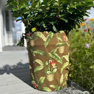 May include: A large, cylindrical plant pot cover with a tropical leaf and flower pattern in shades of brown, green, and red. The cover is filled with a lush green plant, creating a vibrant display for outdoor or indoor use.