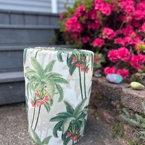 Peut inclure: Un tabouret cylindrique avec un motif de palmiers tropicaux. Le tissu présente des feuilles de palmier vertes et des grappes de fleurs rouges sur un fond beige clair. Le tabouret est à l'extérieur, avec un arrière-plan flou de fleurs roses et d'un escalier gris.