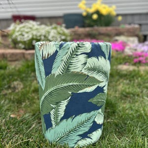 May include: Cylindrical fabric container with a tropical leaf pattern. The design features large green and light green leaves against a dark blue background. The container is set on a bed of green grass.
