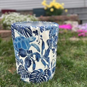 May include: A cylindrical planter with a blue and white tropical pattern featuring turtles and foliage. The planter is made of fabric and sits on a grassy surface, with a blurred background of flowers and a house.