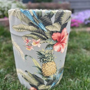 May include: A cylindrical planter with a tropical floral pattern. The design features a pineapple, hibiscus flowers, and a blue parrot against a grey background. The planter is sitting on green grass.