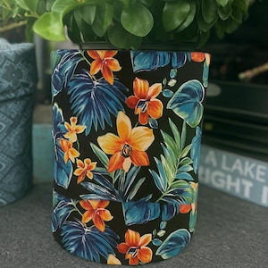以下が含まれることがあります： 鮮やかな熱帯の花柄の円筒形のプランター。オレンジと黄色の花、青い葉、緑の葉が黒い背景にデザインされています。プランターには緑の葉の植物が入っています。