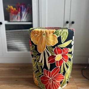 May include: A cylindrical fabric storage container with a vibrant floral pattern. The design features large orange, yellow, and red flowers with green leaves against a black background. The container sits on a wooden floor.