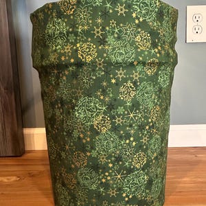 May include: A cylindrical green fabric plant pot cover with a repeating pattern of gold and white snowflakes and floral designs. The cover is placed over a plant pot, adding a decorative touch to the plant.